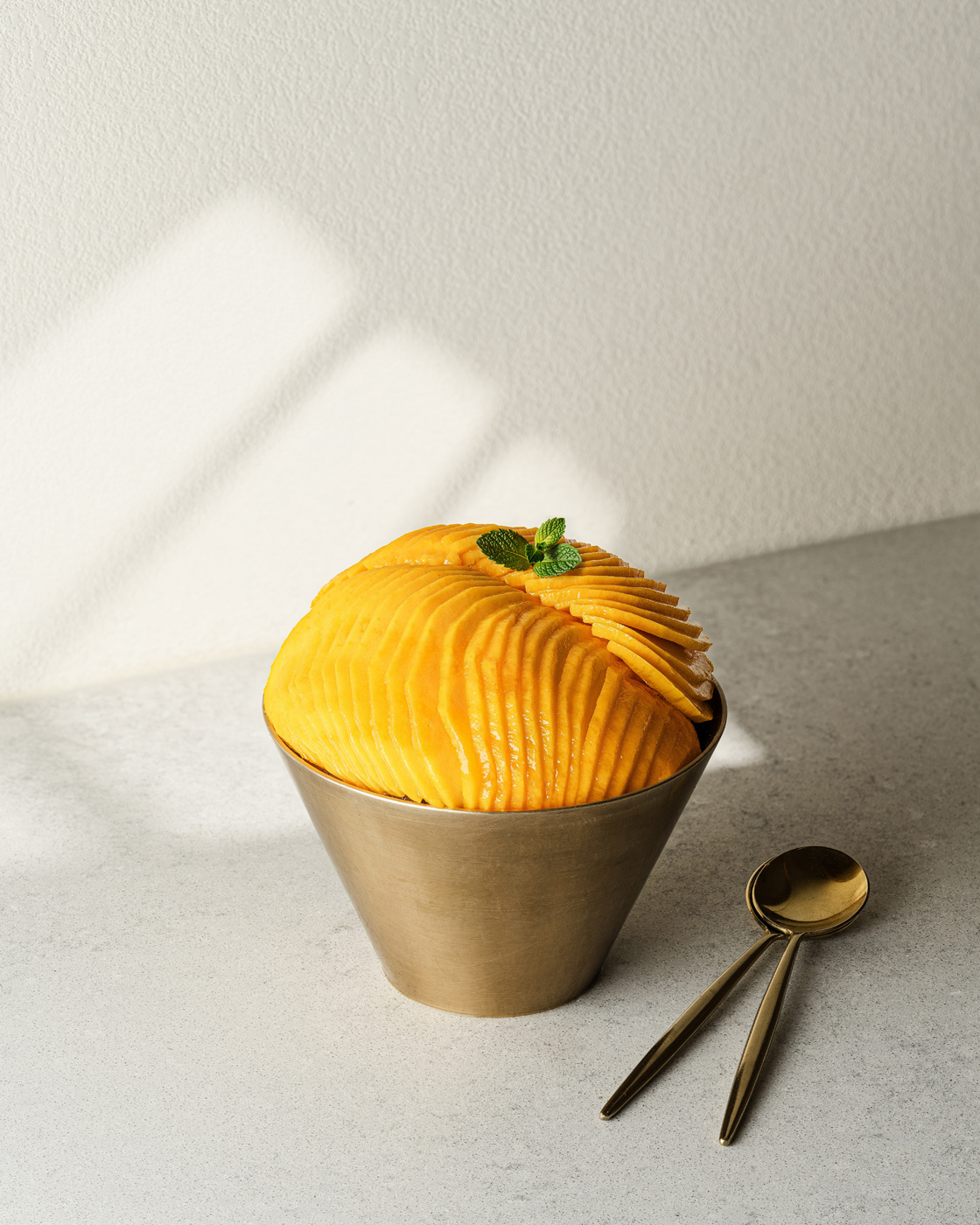 A meticulously sliced mango arranged in an elegant pattern within a sleek, gold-toned cup set against a minimalist gray backdrop. The lighting creates a soft, ambient mood highlighting the fruit's vibrant color and texture, accompanied by a pair of golden spoons.