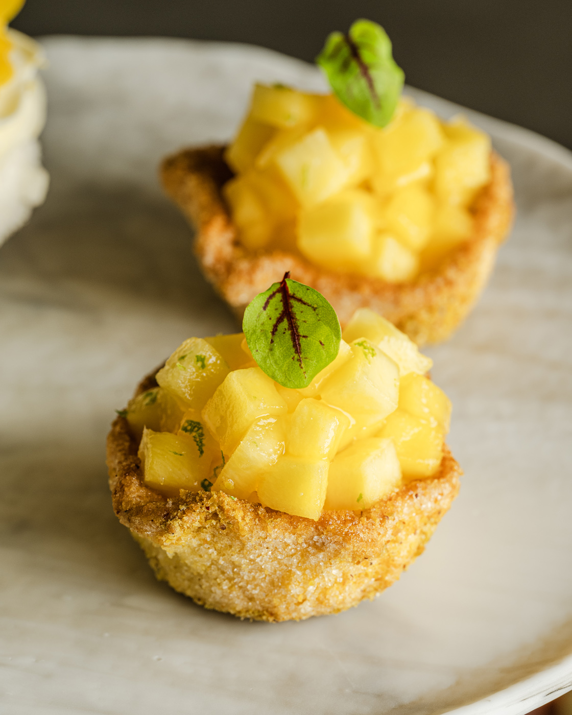 Close-up of gourmet pineapple tartlets, elegantly arranged on a marble plate. The tartlets are topped with vibrant yellow pineapple cubes and a delicate green leaf garnish, captured in a soft, ambient light enhancing their rich textures and inviting colors.