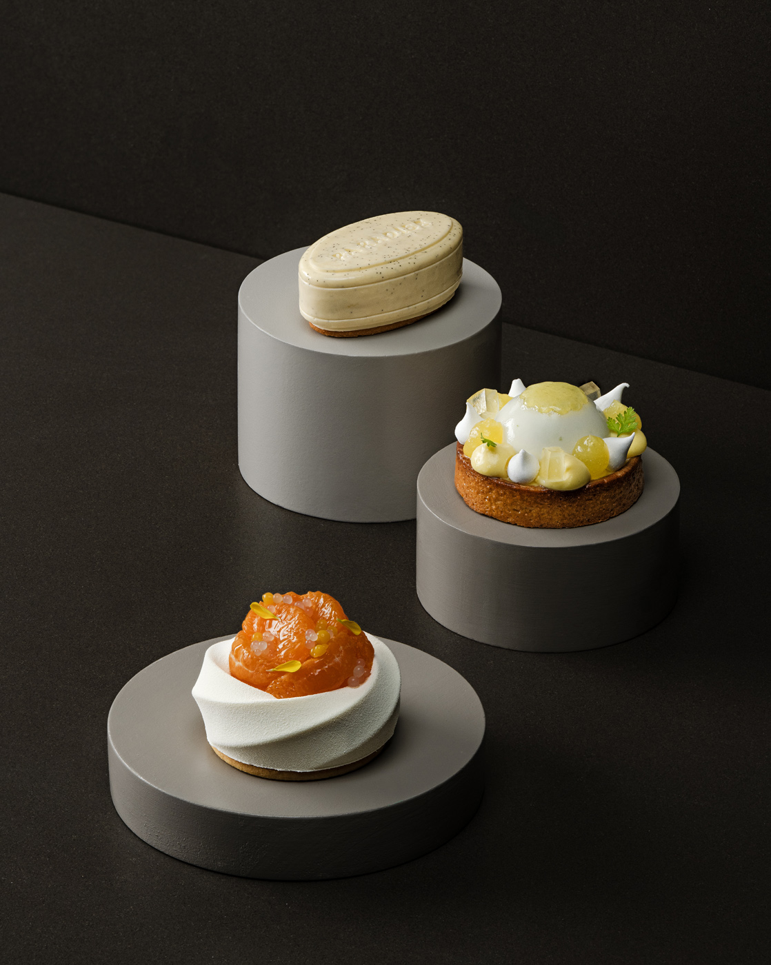 A sophisticated display of three gourmet desserts placed on minimalist grey pedestals against a dark background. The desserts include a vanilla-glazed elongated cake, a citrus tart with delicate white meringue peaks, and a modern dessert with a vibrant orange topping, each artfully arranged and highlighted by soft, even lighting.