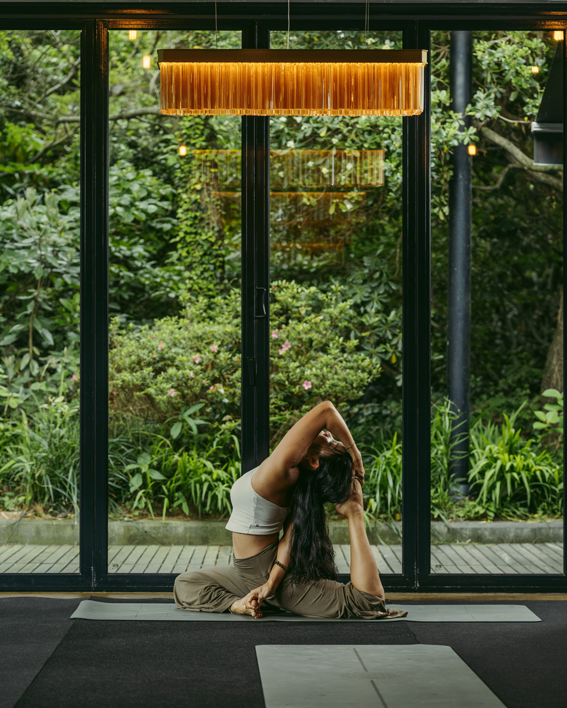 A woman practicing an elegant yoga pose indoors, framed by large glass windows revealing a lush, green garden outside. The ambient lighting creates a calming and serene atmosphere, highlighting the flexibility and grace of the pose.
