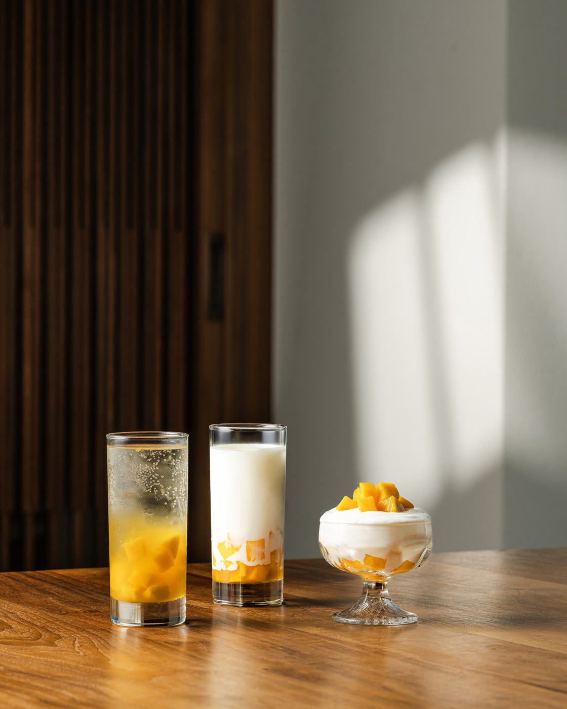 Three glass containers on a wooden table, featuring a mango dessert and drinks, bathed in soft, natural lighting with warm, inviting tones.