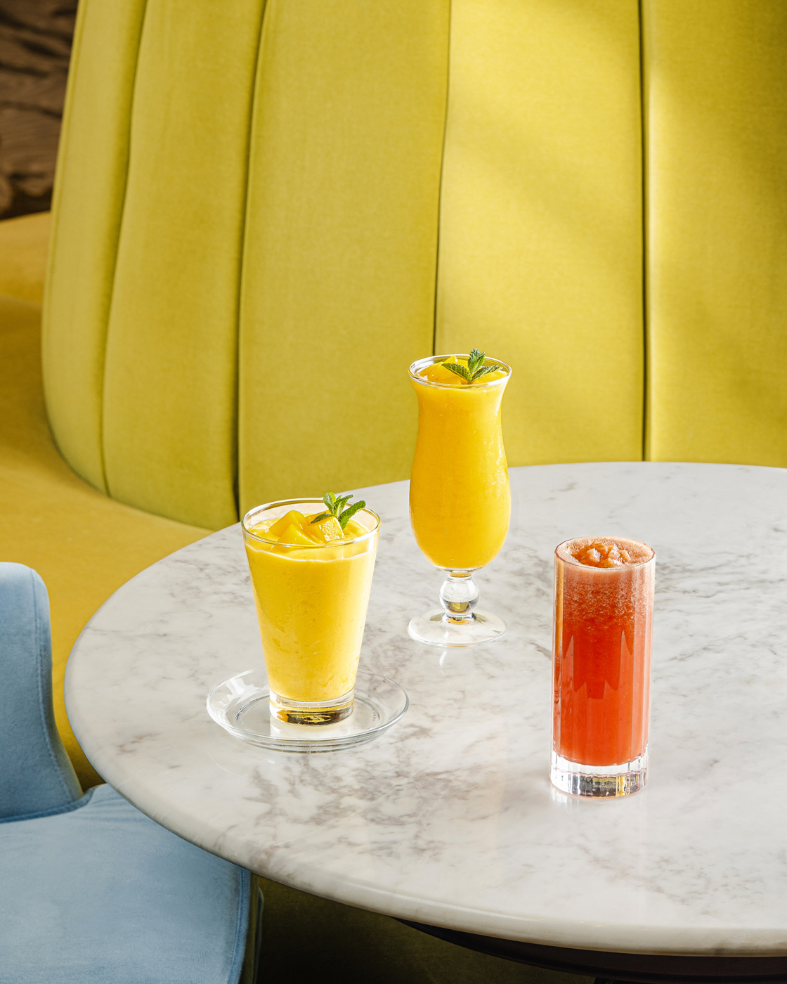 Three refreshing drinks displayed on a marble table: two tropical orange smoothies in different glass styles, both garnished with mint leaves, and a vibrant red juice served in a tall clear glass. The setting is enhanced by warm, ambient lighting and a sophisticated mustard-colored backdrop.