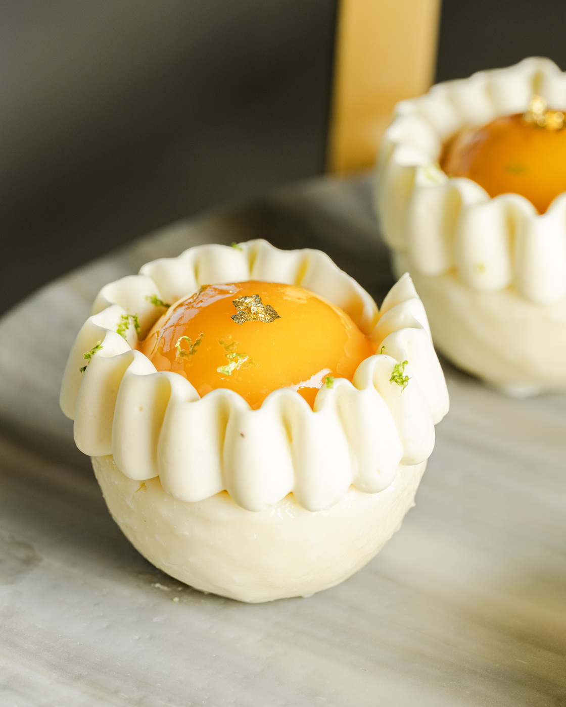 A close-up view of a delicate citrus mousse dessert topped with a glossy orange glaze and a touch of golden leaf, surrounded by piped white cream. The dessert is artistically displayed on a marble surface, with soft lighting accentuating the creamy textures and vibrant colors.