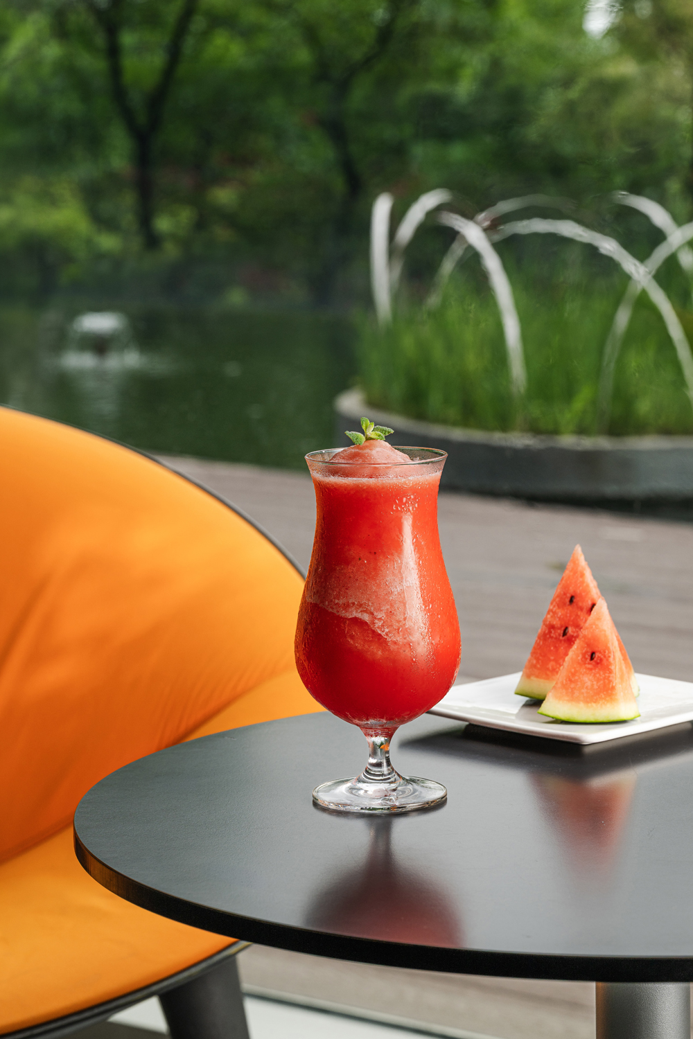 A vibrant red watermelon cocktail in a curvaceous glass, garnished with mint, placed on a sleek black table beside watermelon slices. The background features a serene pond and a bright orange chair, creating a lively and refreshing atmosphere.