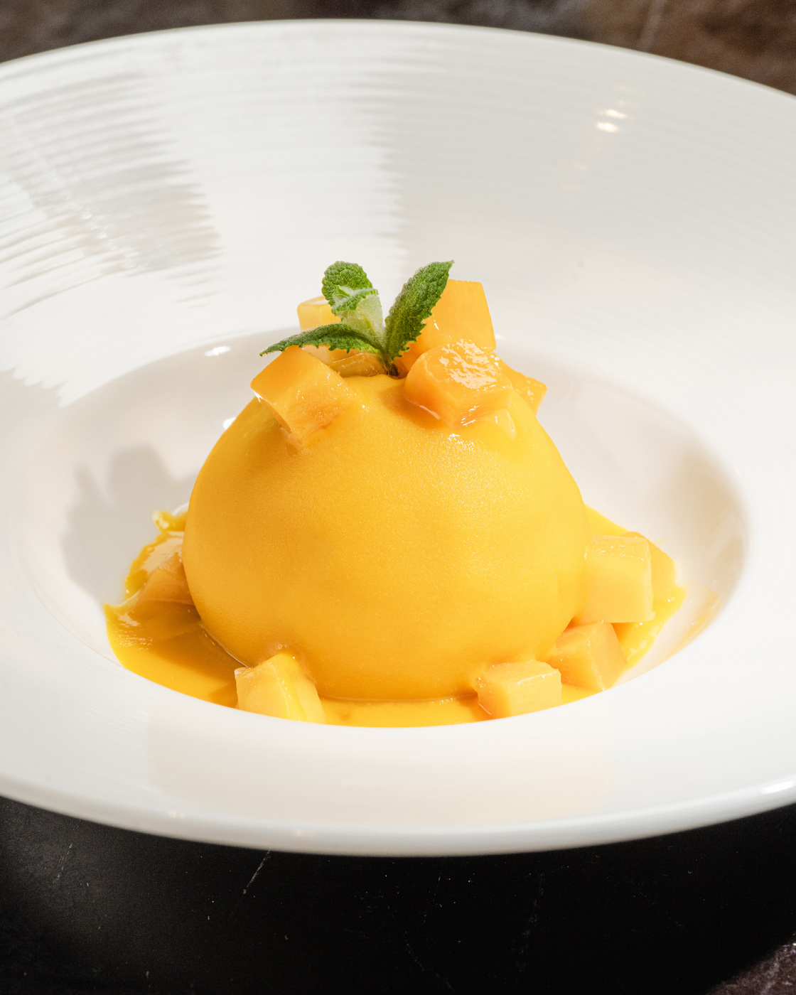 Elegantly presented dome-shaped mango mousse dessert garnished with fresh mint and diced mango, set on a white plate with soft, natural lighting highlighting its silky texture.