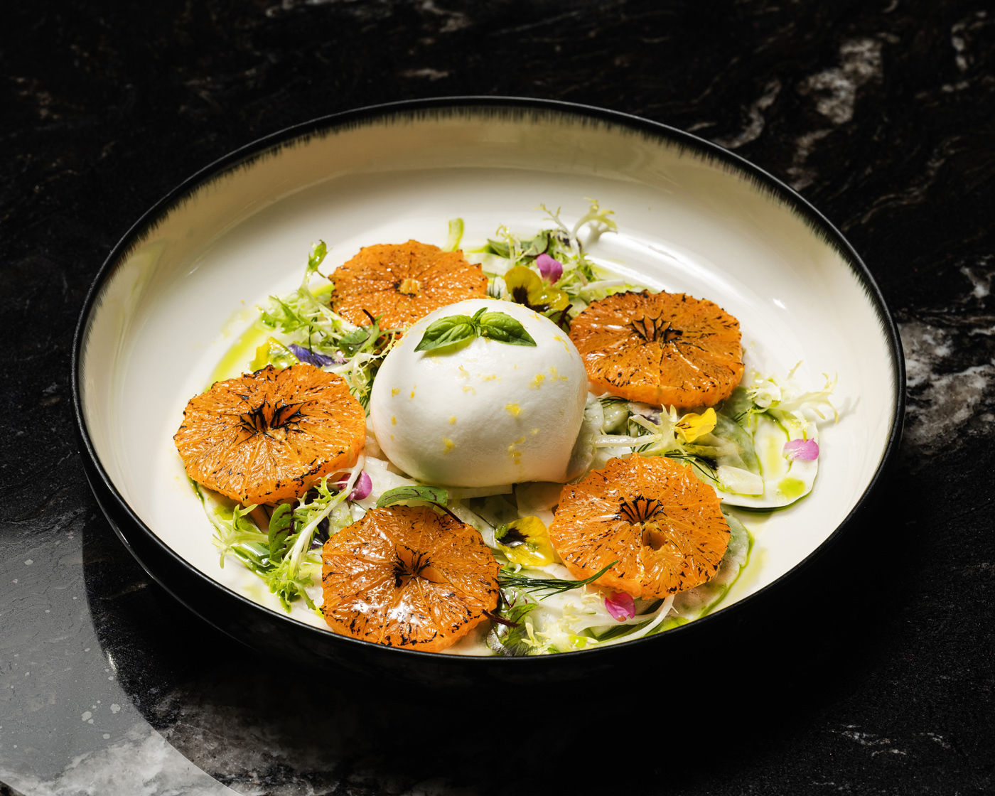A visually striking dish featuring a creamy burrata cheese centerpiece surrounded by grilled orange slices atop fresh greens, presented in a white bowl against a marble surface with chiaroscuro lighting enhancing texture and color.