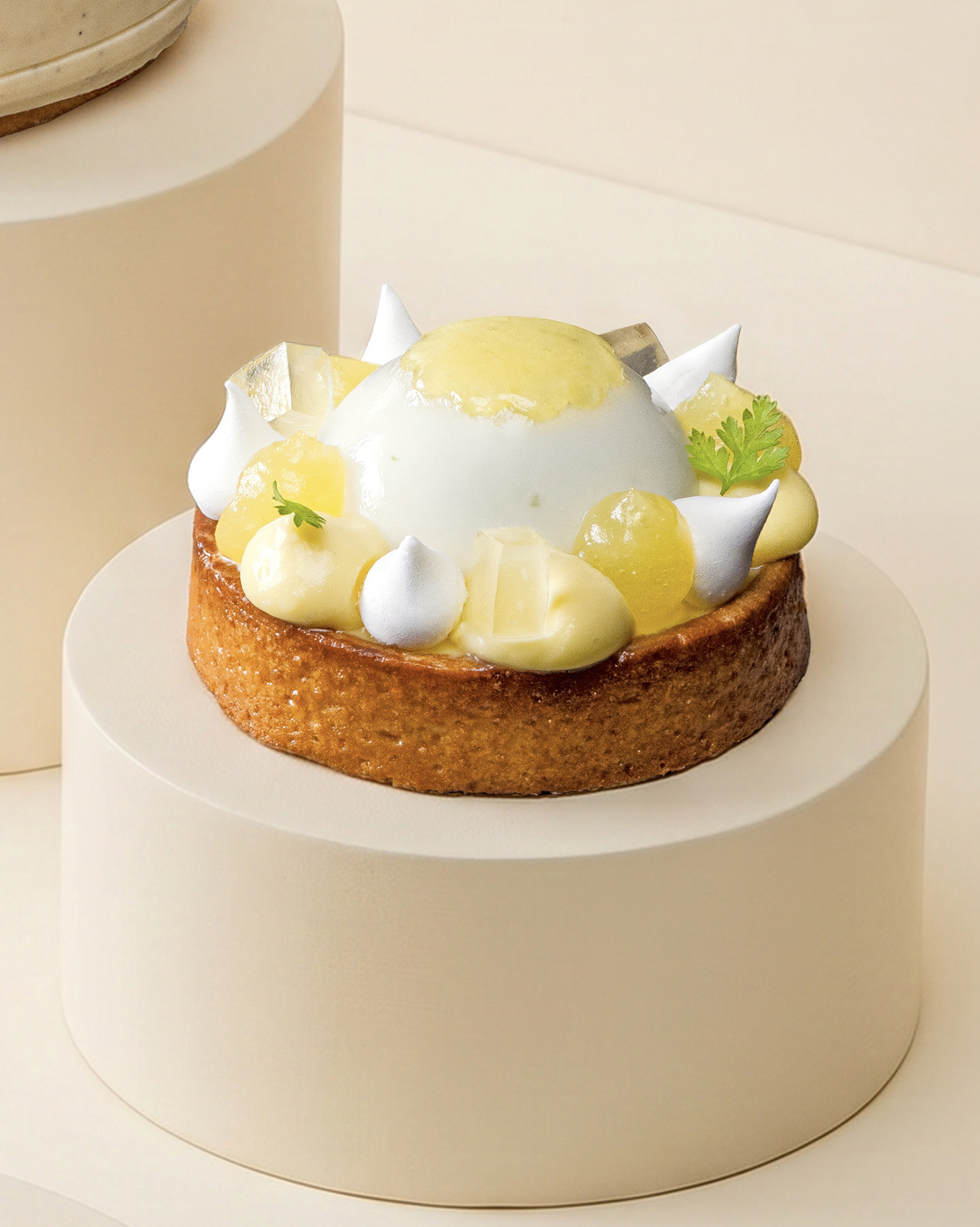 A lemon meringue tart is artistically presented on a cream-colored pedestal. The tart features a glossy lemon dome, surrounded by dollops of white meringue and lemon curd, with a delicate piece of candied citrus and small herb leaves for garnish. The bright dessert is contrasted against a neutral background, enhancing its sophisticated and delightful appearance.