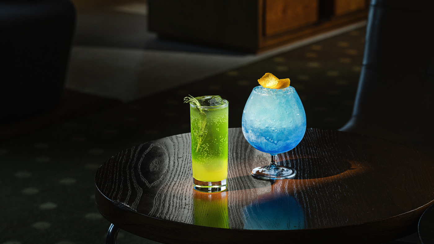 Two exquisitely crafted cocktails on a dark wooden table; a vibrant green drink with rosemary garnish in a tall glass and a striking blue cocktail with orange peel in a rounded glass, illuminated by gentle ambient lighting.