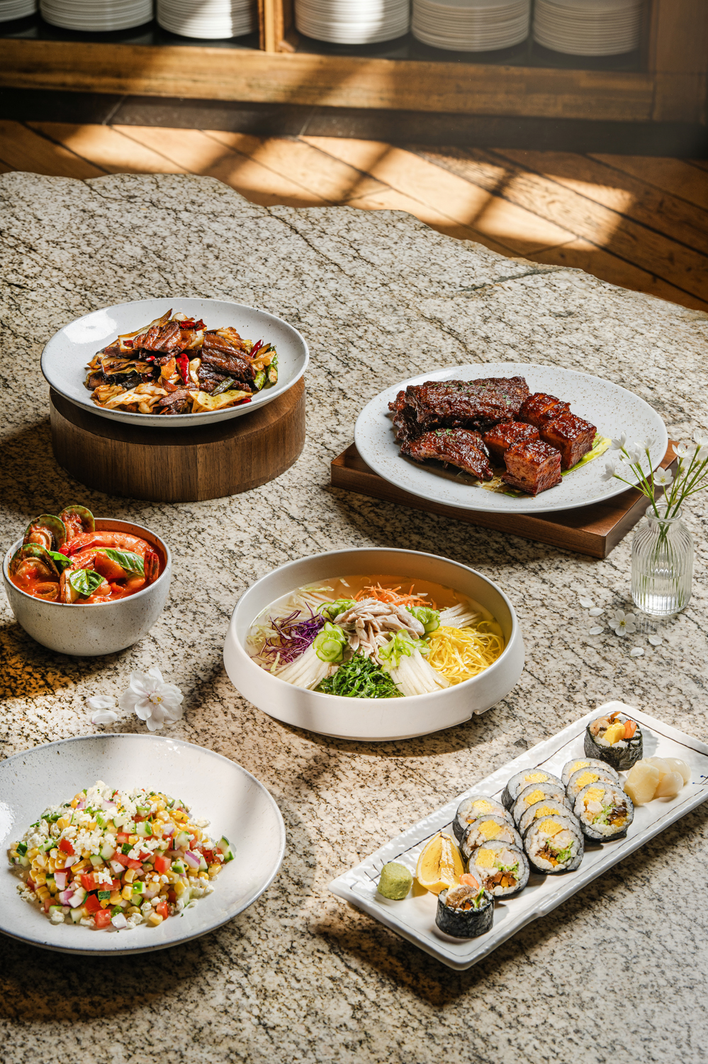A sophisticated spread of Korean dishes including grilled meat, assorted vegetables, noodles, kimbap, and salad, artfully arranged on a marble table under soft daylight.