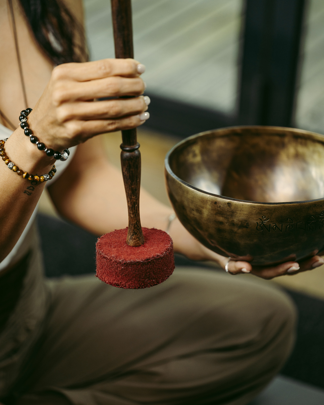 A close-up of a person's hands holding a Tibetan singing bowl and mallet, with soft ambient lighting and a peaceful atmosphere. The person's wrist is adorned with beaded bracelets, enhancing the calming setting.