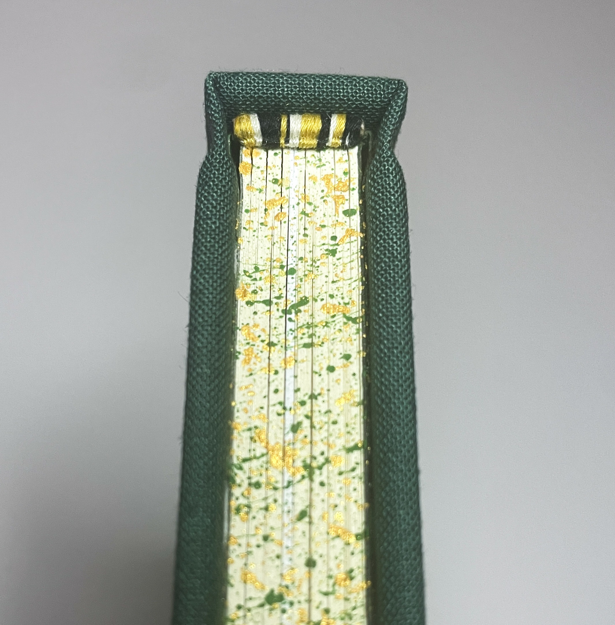 The top of a hand-bound book, focusing on the endbands, which are French double-core and sewn with black, white, and gold thread. The edges are speckled with green and gold paint.
