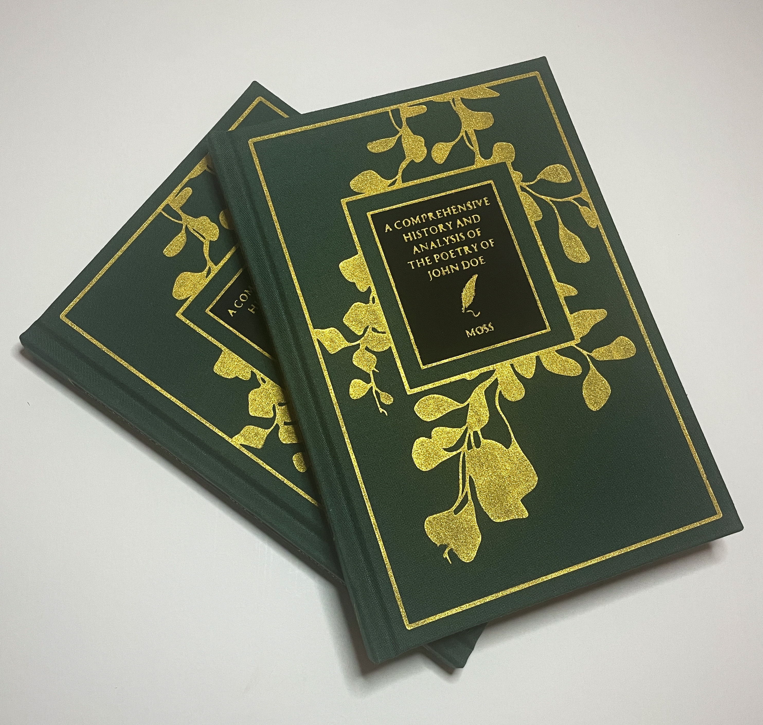 Two identical hand-bound books stacked atop one another. They are bound in dark green cloth with a gold-painted vine pattern on the cover that surrounds a black rectangle with the title, 'A Comprehensive History and Analysis of the Poetry of John Doe,' and author name, 'Moss,' painted atop it in gold.