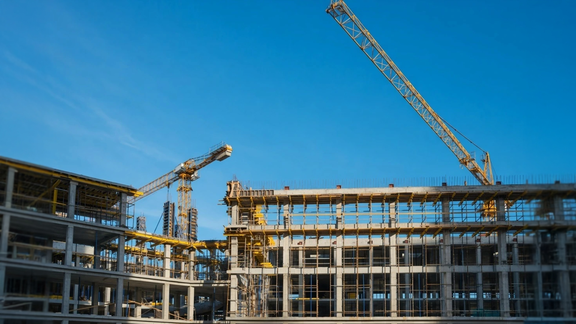 Construction site with unfinished concrete buildings, scaffolding, and two yellow tower cranes under a clear blue sky.