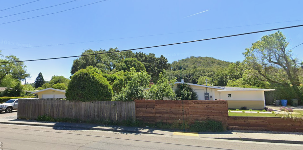 Existing ranch house behind a wood fence, with trees beyond