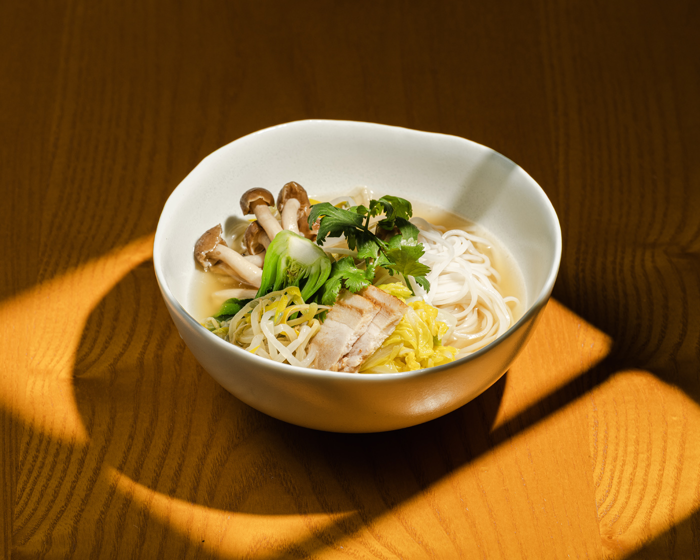 A beautifully composed bowl of noodle soup featuring slices of tender pork, fresh mushrooms, and vibrant greens, set against a warm wooden background illuminated by soft, natural lighting. The composition highlights the rich textures and fresh ingredients in a flambient mood.