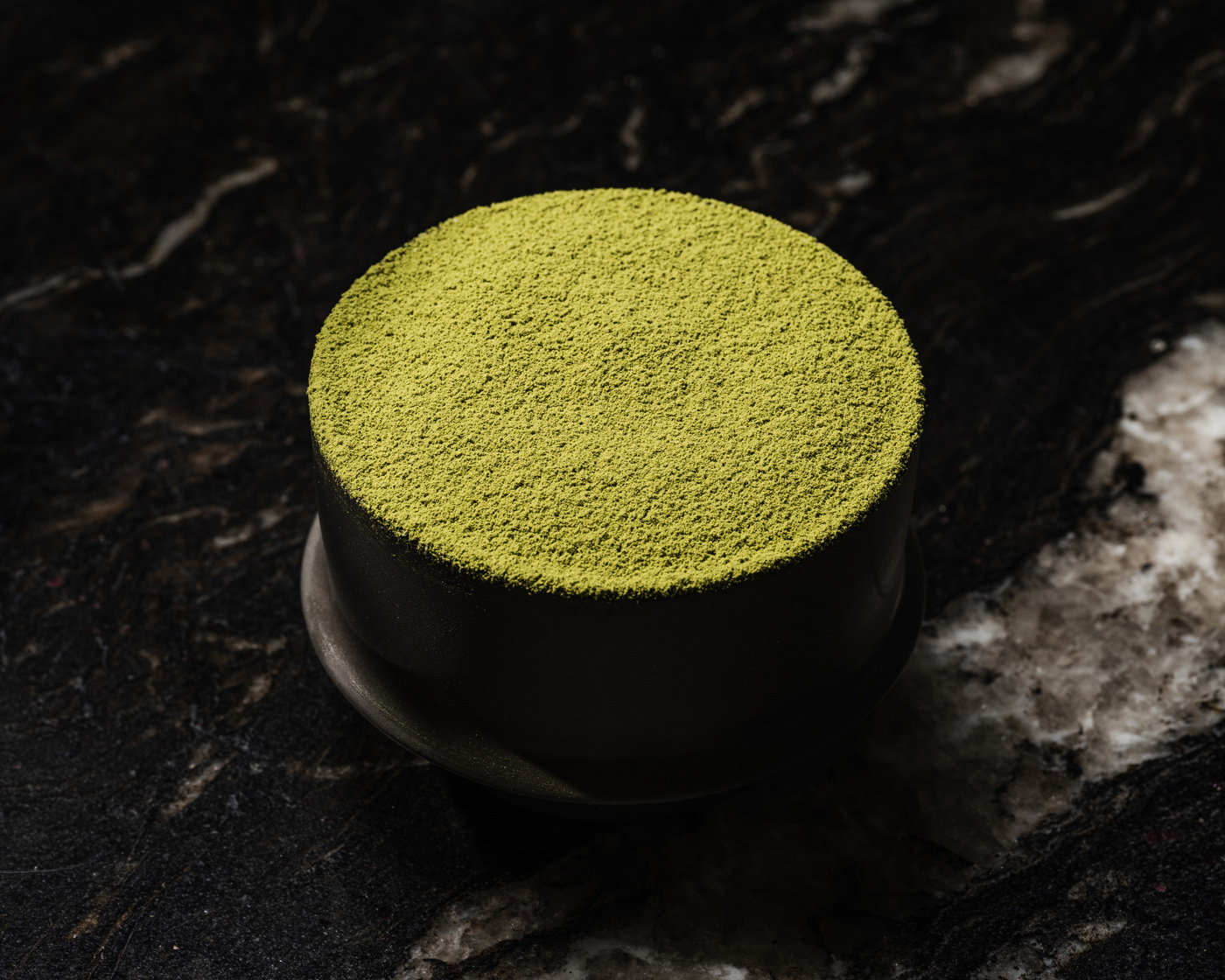 A sophisticated dessert topped with vibrant matcha powder, presented on a dark marble surface. The image captures the fine-textured surface of the dessert under subtle lighting, creating a luxurious and serene atmosphere.