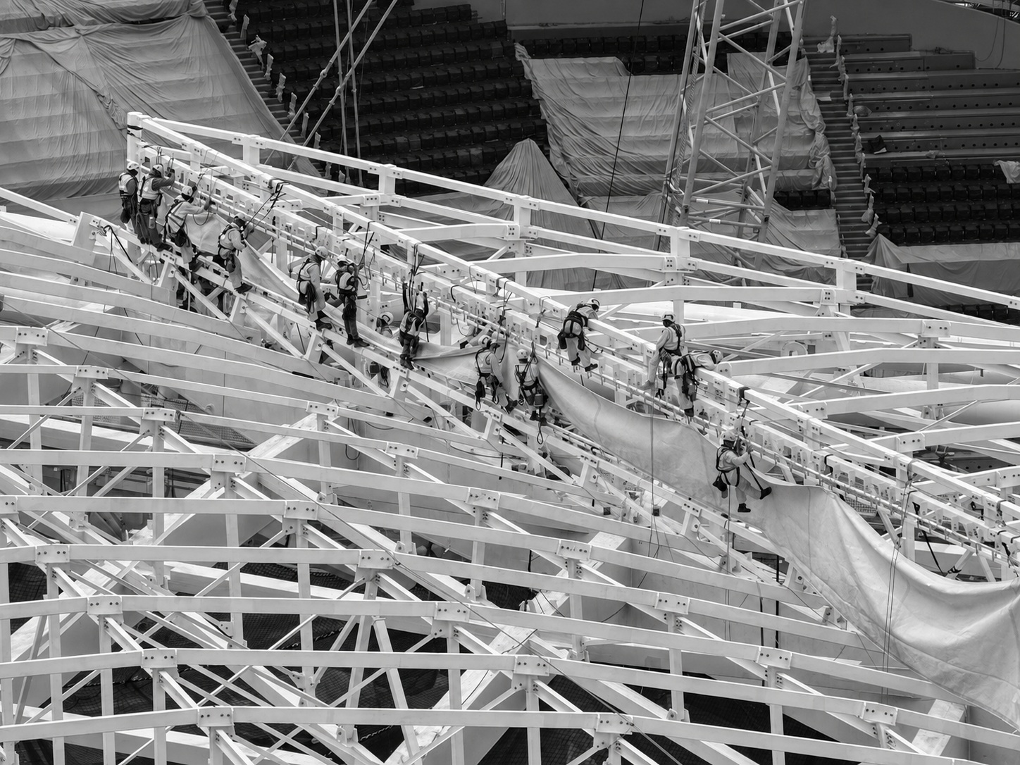 Rope access technicians in harnesses installing a tensile membrane panel across a stadium roof truss