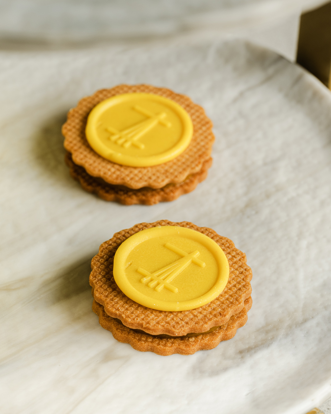 Close-up image of two gourmet lemon cookies with a decorative yellow icing on a beige plate, softly lit, showcasing detailed textures and a minimalist composition.
