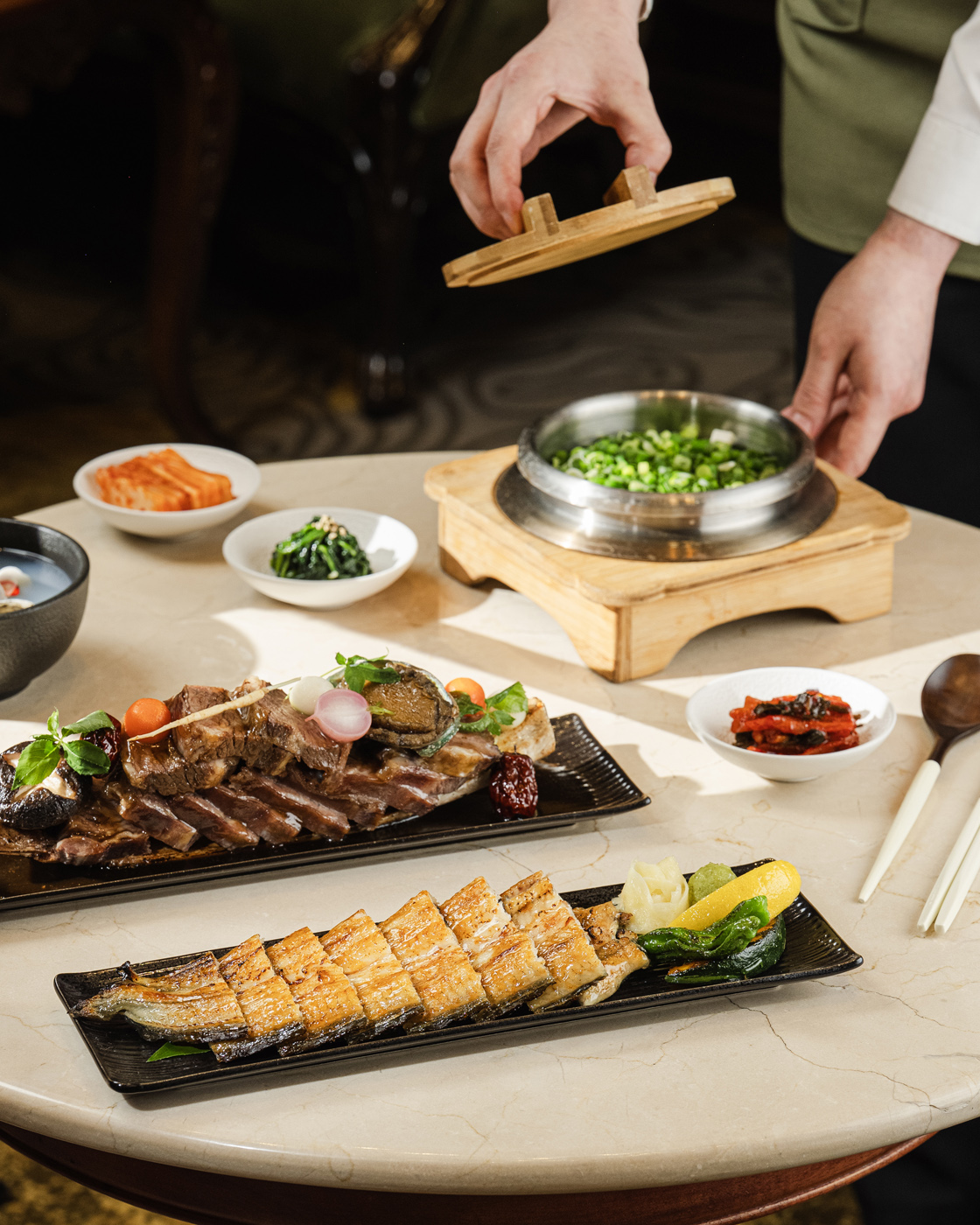 A luxurious Korean meal arrangement on a marble table, featuring grilled eel and beef ribs on black plates, accompanied by a traditional stone pot rice with green onions. The lighting is bright and natural, highlighting the rich textures and colors of the dishes.