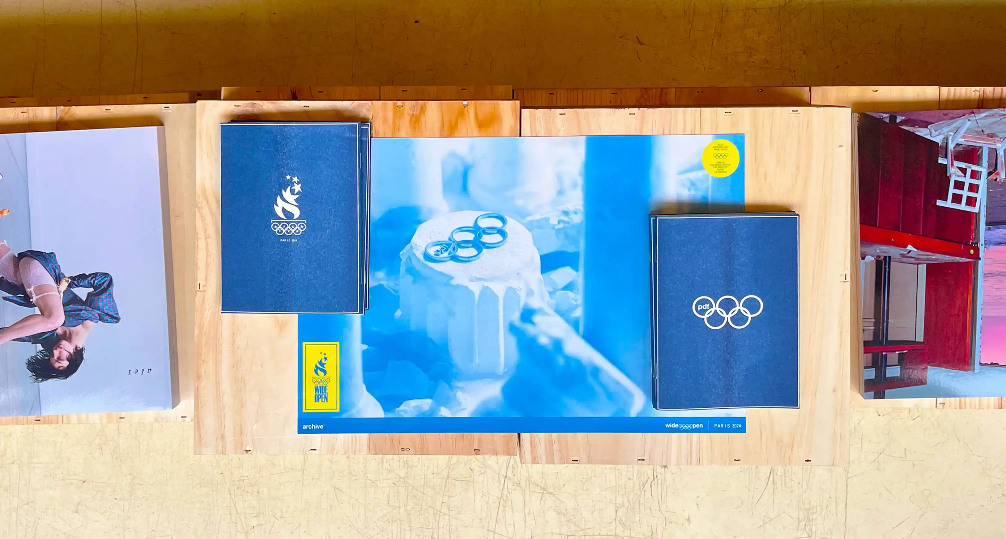 Archive PDF wide open Paris 2024 print publication spread laid out on a wooden surface featuring the Paris Olympics flame logo zine the pdf Olympic rings cover and a blue toned editorial poster